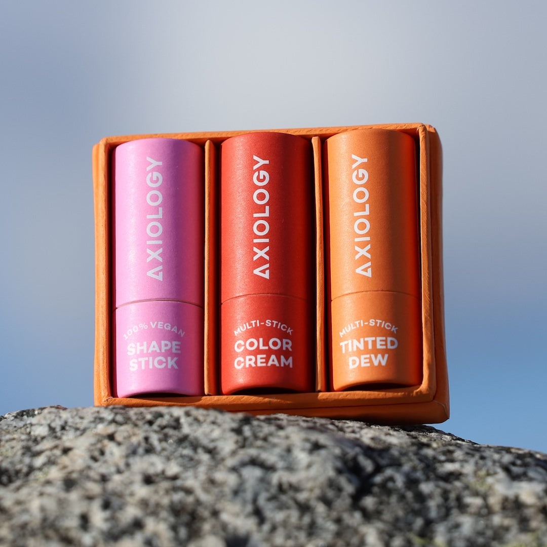 Three Axiology makeup sticks in a case on a rock with a blurred natural background