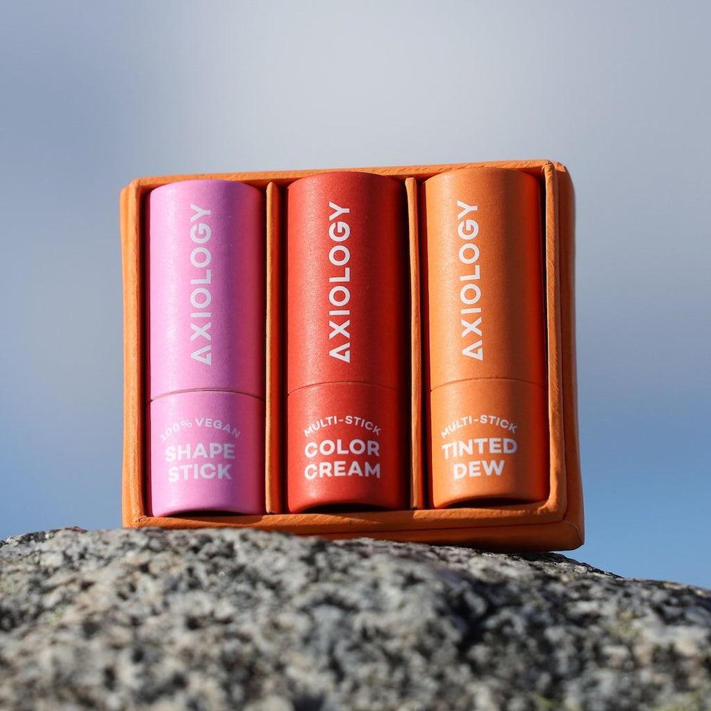 Three Axiology makeup sticks in a wooden case on a rock with a blurred natural background