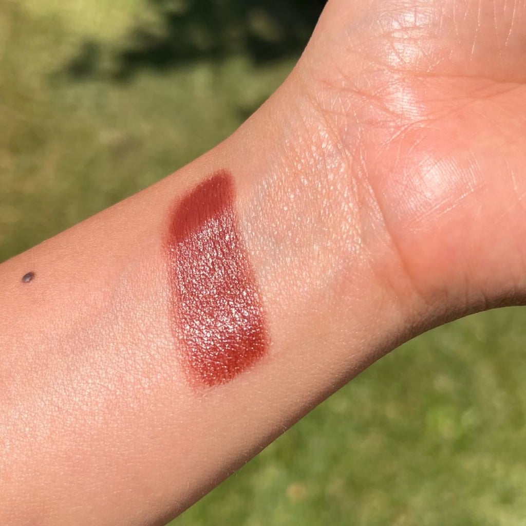 Brick red lipstick swatch on a person's arm with a blurred green outdoor background