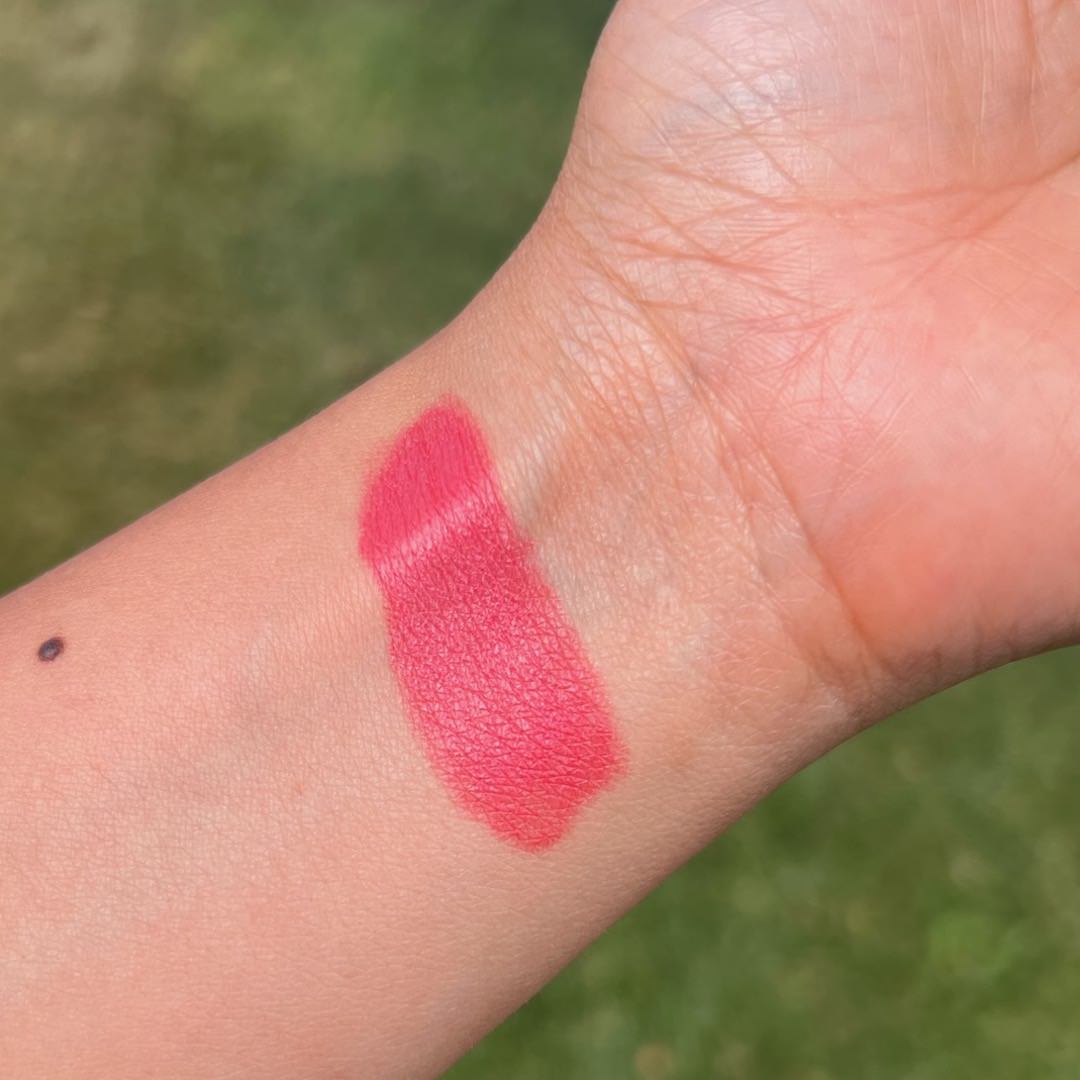 Pink lipstick swatch on a person's arm with a blurred green background