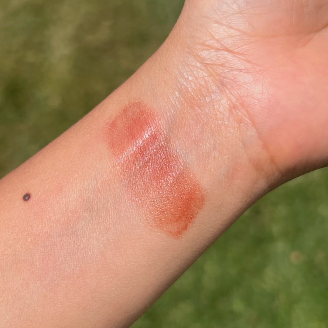 Close-up of a swatch of makeup on a person's arm with a green outdoor background
