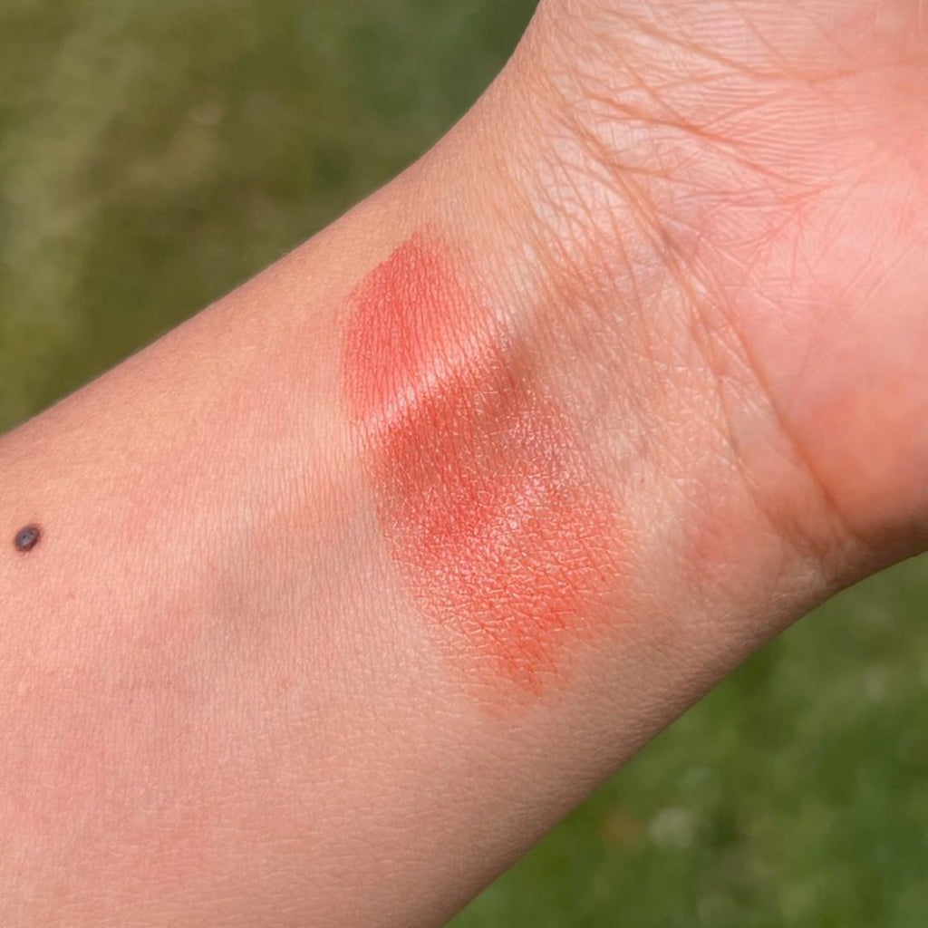 Close-up of a swatch of coral-colored makeup on a person's wrist with a blurred green background.