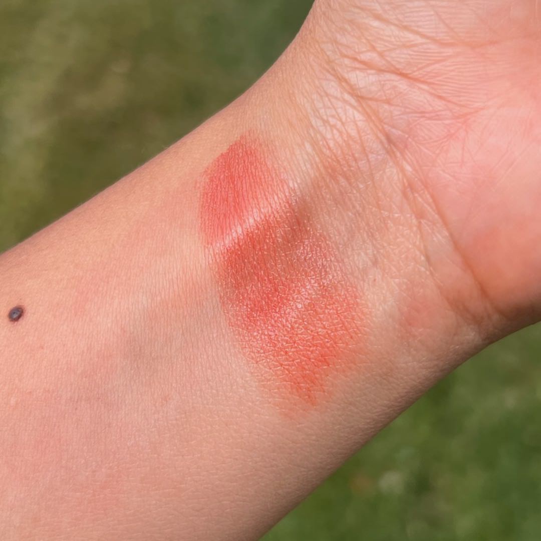 Close-up of a swatch of coral-colored makeup on a person's wrist with a blurred green background.