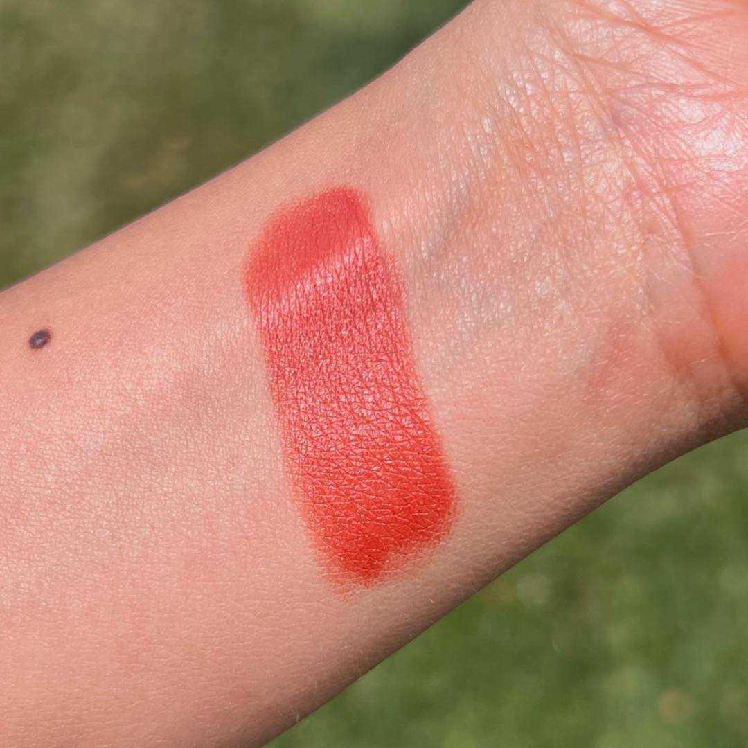 Orange red lipstick swatch on a person's arm with a blurred green background
