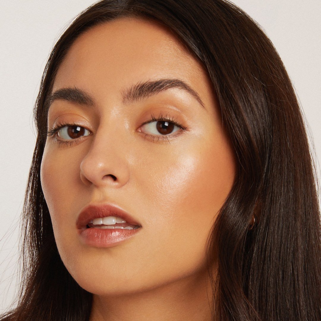 Close-up of a woman with a neutral expression wearing foundation makeup on a plain background