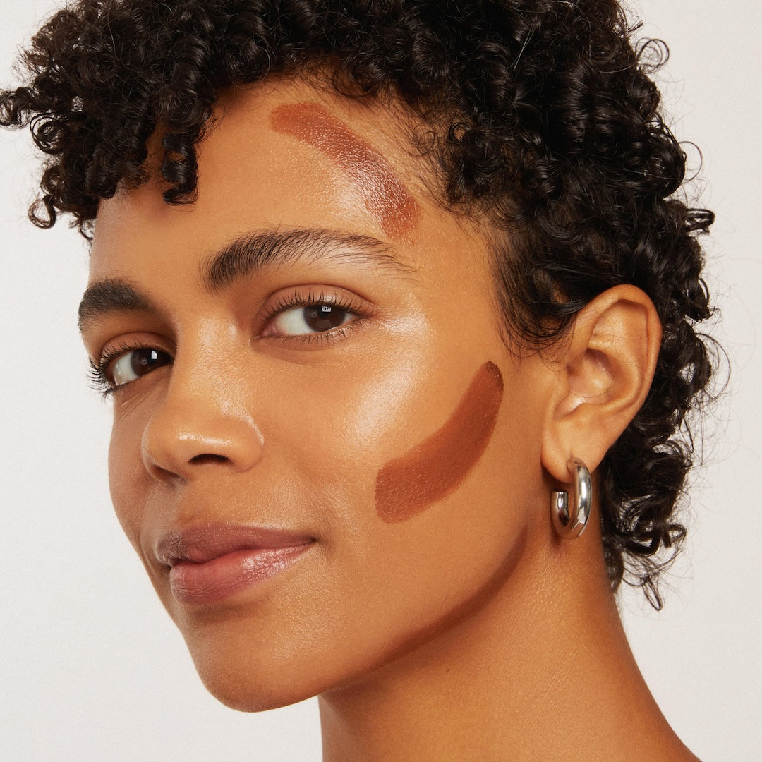 Close-up of a woman's face with makeup swatches on her cheek, against a neutral background