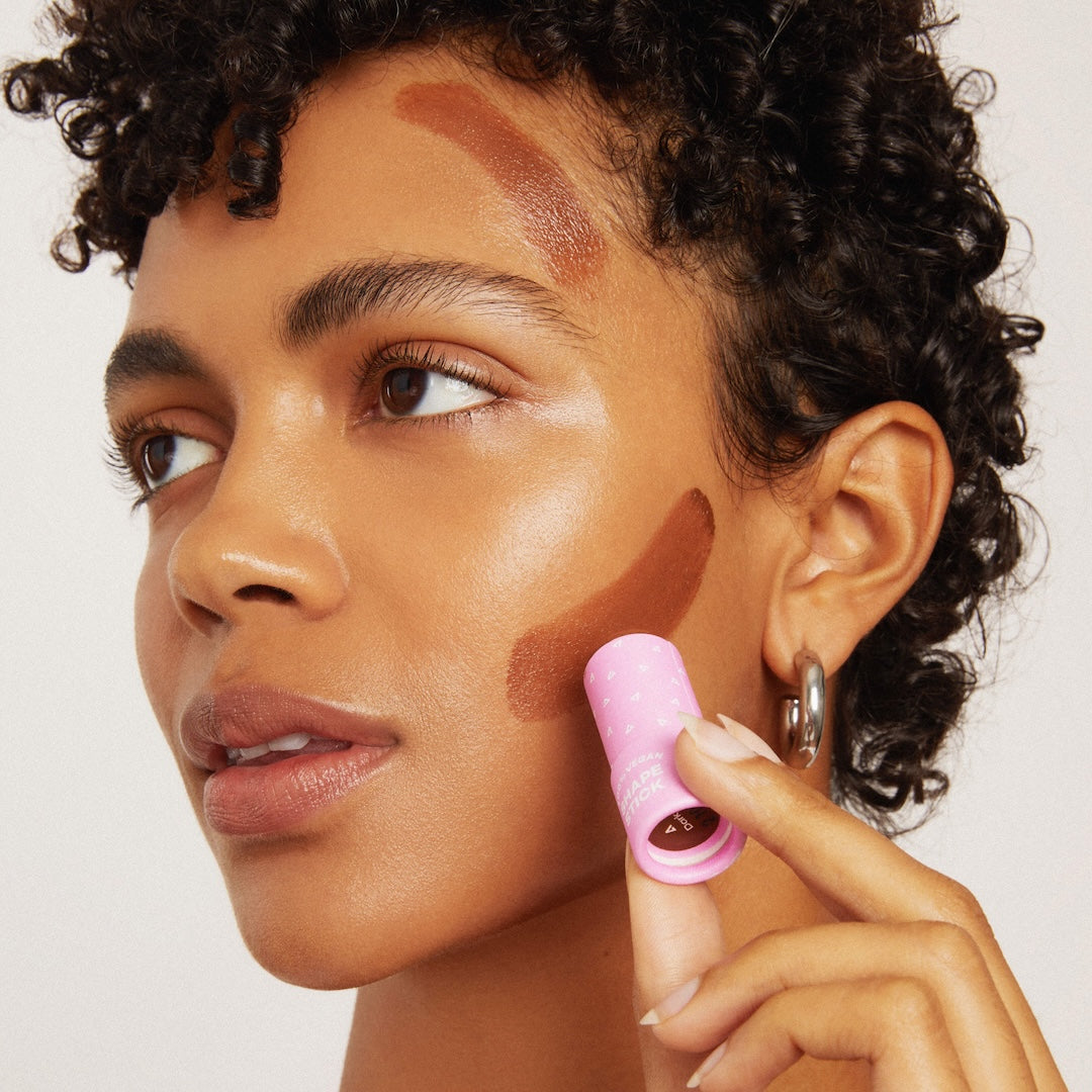 Woman applying makeup with a pink tube applicator on a neutral background