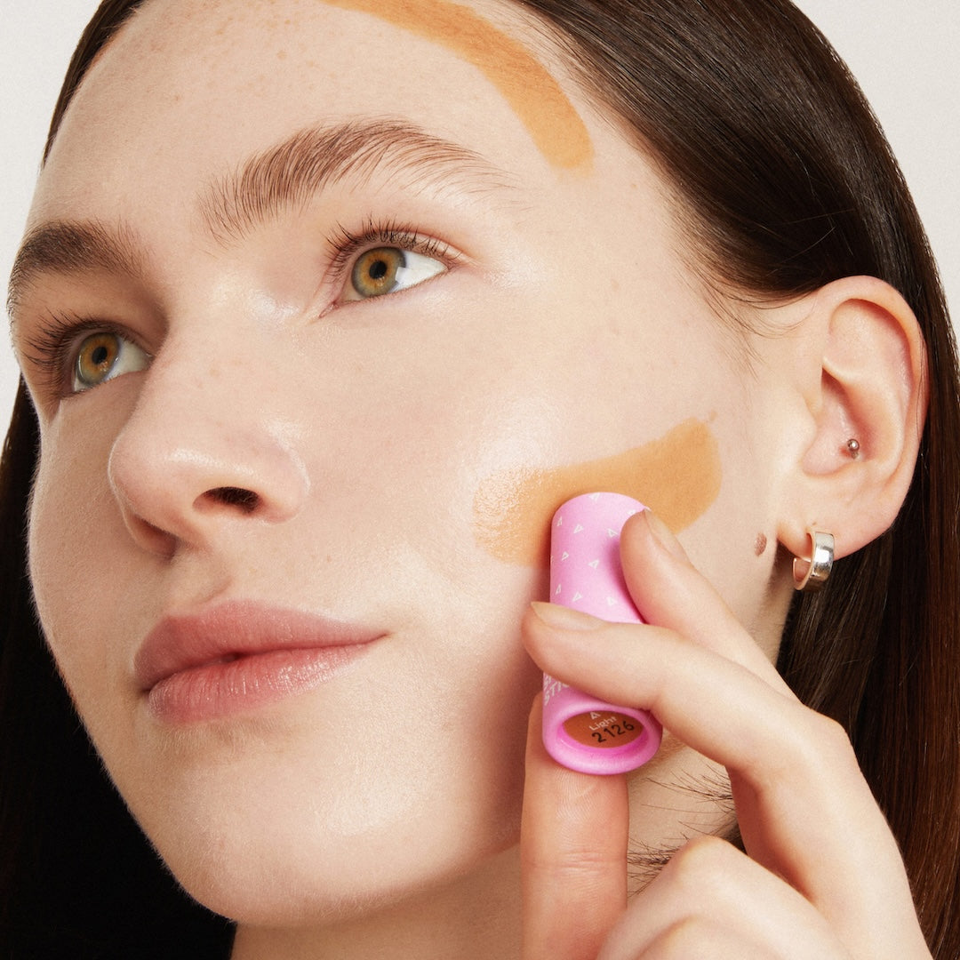 Woman applying makeup with a pink tube applicator on a neutral background
