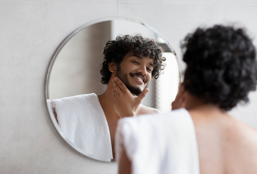 Man in a bathroom looking at himself in the mirror, touching his beard.