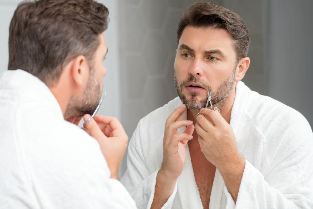 Man in a white robe trimming his beard with small scissors in front of a mirror.
