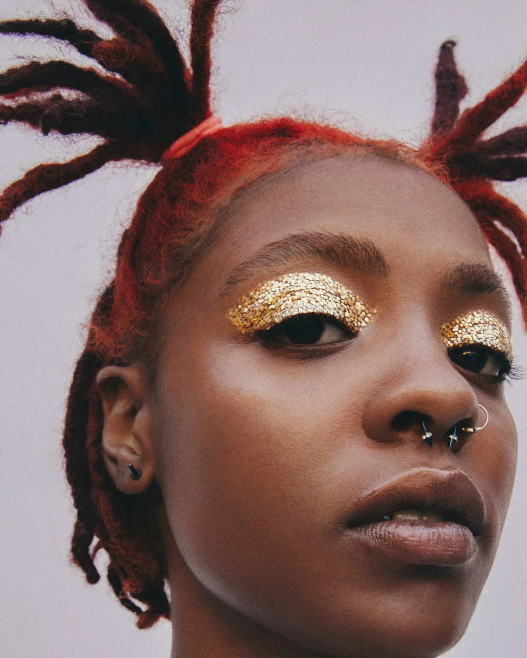 Close-up of a person with red hair styled in braids, wearing gold glitter eye makeup on a plain background.